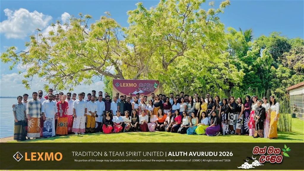 Large group of people posing outdoors under a tree at a Lexmo event, banner in background reading LEXMO and Alluth Avurudu 2026 read aloud later text overlaps bottom bar to right: Tradion & Team Spirit United.