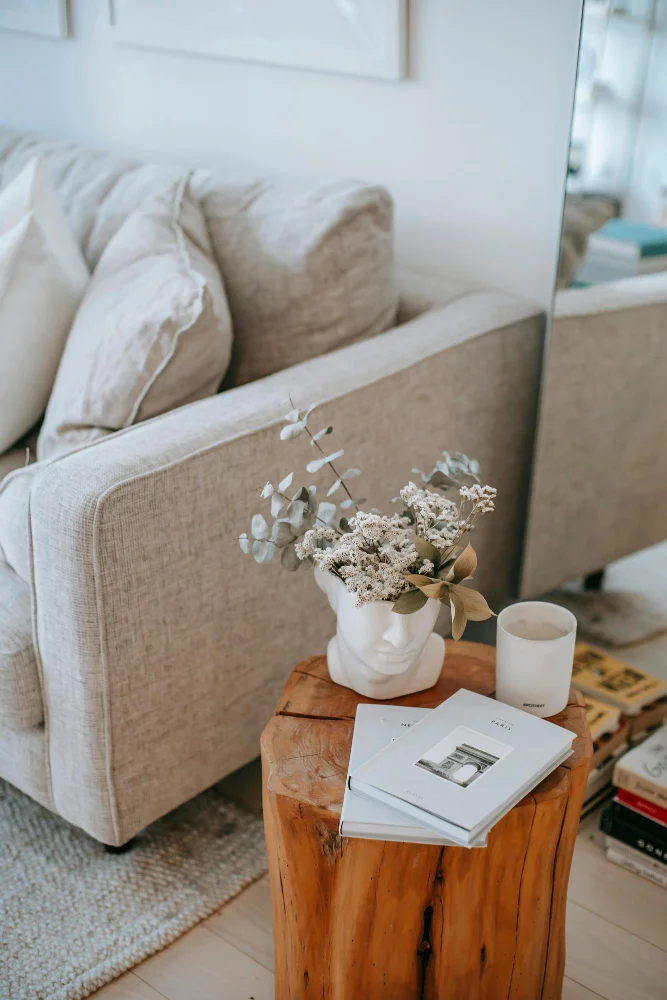 Beige sofa in a cozy living room with a wooden stump side table holding a white vase with dried flowers, a white candle, and a couple of books.