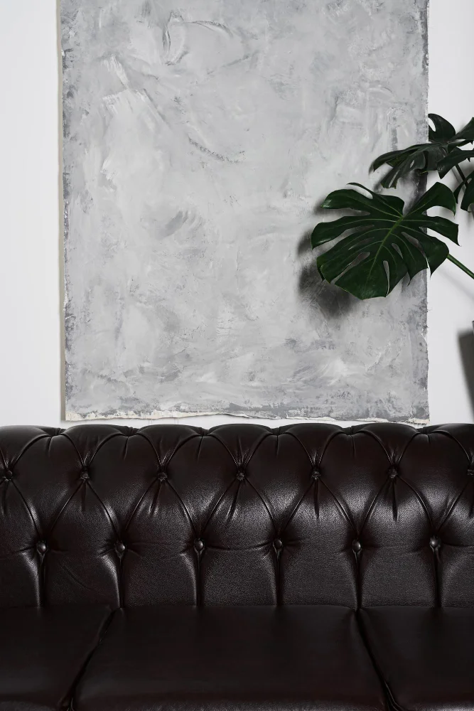 Grey abstract painting mounted on a white wall above a dark tufted leather sofa, with a Monstera leaf peeking in from the right side.