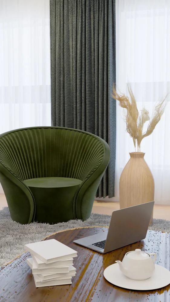 Green curved lounge chair beside a wooden coffee table with an open laptop, a teapot on a tray, and a stack of white papers; background has a vase with dried grasses and sheer curtains.