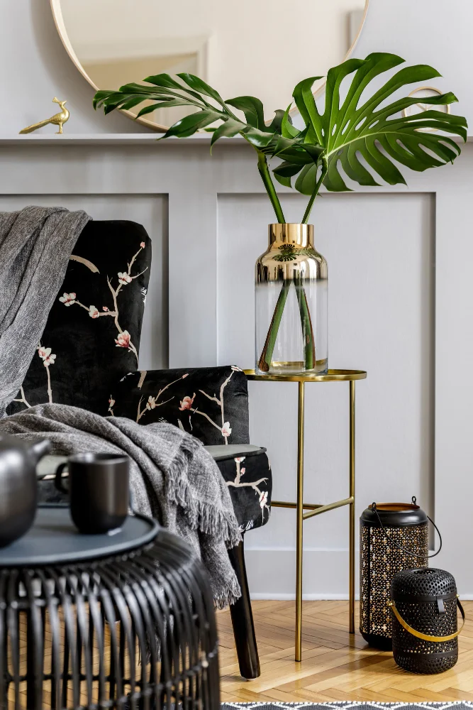 Cozy seating area with a black floral chair, gray throw, a gold-side table holding a glass vase with a Monstera leaf, and lanterns nearby.