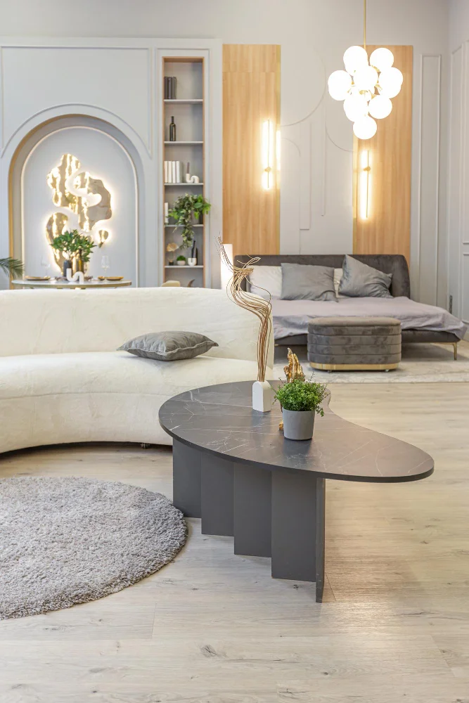 Modern living room with a curved white sofa, dark oval marble coffee table, and a gray circular rug; decorative sculpture and potted plant on the table. In the background, a gray sofa with cushions, built-in shelving, and warm vertical lighting against wooden panels.